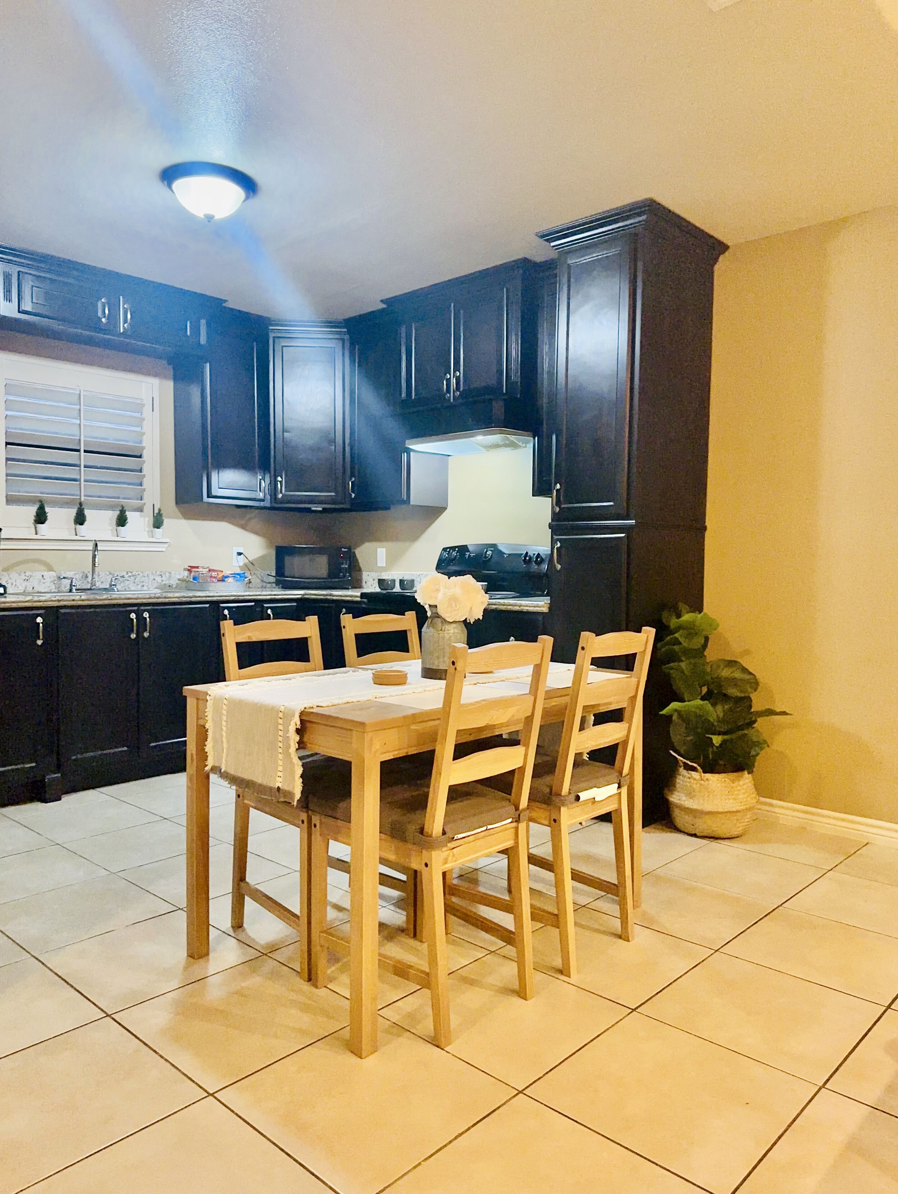 A kitchen with dark cabinets, a wooden table, and chairs. There is a potted plant and a vase with flowers on the table.