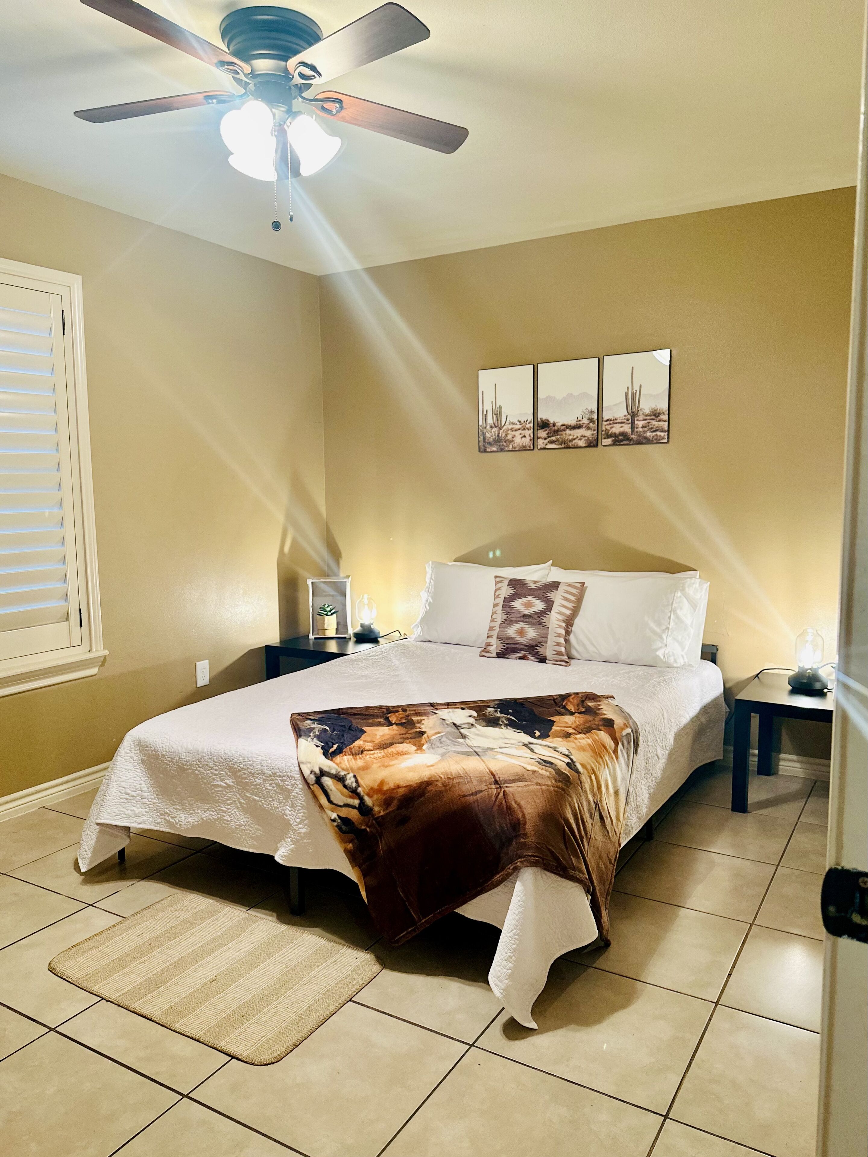A bedroom with a bed, a ceiling fan, a nightstand, a wall-mounted picture, and a window with blinds.