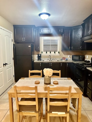 A kitchen with dark wooden cabinets, a black refrigerator, a microwave, and a stove. There is a dining table with chairs and a vase with flowers on it.