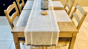 Une table à manger en bois, recouverte d’une nappe tissée, six chaises en bois et un vase rempli de fleurs blanches.