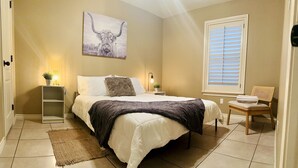 Une chambre à coucher comprenant un lit, une table de chevet, une chaise, une fenêtre avec des stores et un tableau accroché au mur.