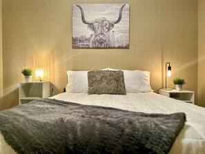 A bedroom with a bed, pillows, a blanket, a wall-mounted picture of a bull, bedside tables with lamps, and potted plants.