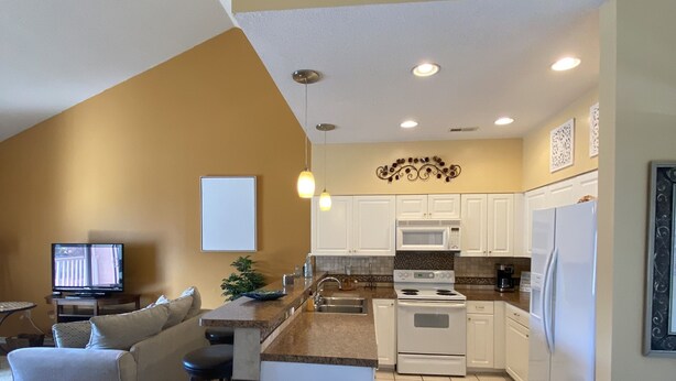 A modern kitchen with white cabinets, a stainless steel refrigerator, and a built-in oven. There is a dining area with a table and chairs, and a living room with a sofa and a TV.