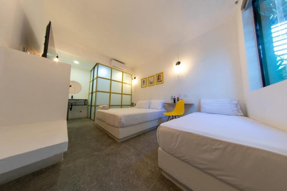 Habitación de hotel compacta con dos camas individuales, un escritorio pequeño, una silla y una ventana con persianas.