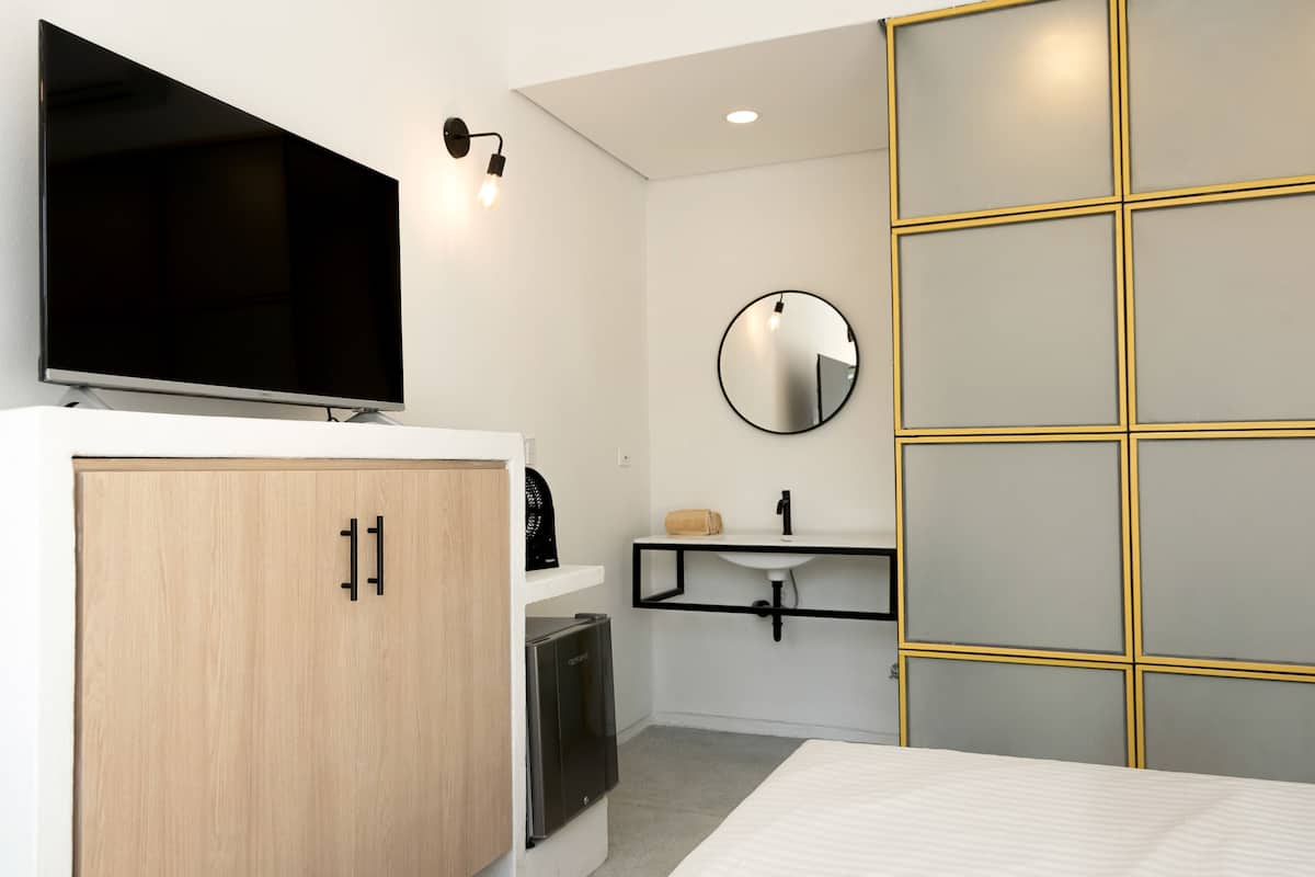 A modern hotel room with a flat-screen TV, a wooden wardrobe, a small refrigerator, a bedside table, a round mirror, and a built-in cabinet with a gold frame.