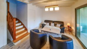 Ett modernt vardagsrum med en soffa, ett soffbord och två svarta pallar. En trätrappa syns till vänster och en glasdörr till höger.
