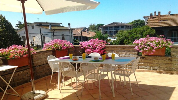 Une terrasse avec une table et des chaises, entourée de plantes en pot aux fleurs roses et d’un mur de pierre.
