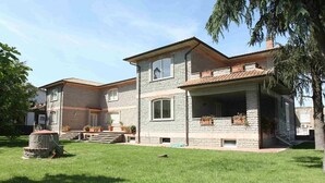 Une maison en pierre à deux étages, dotée d’un porche couvert, d’un puits en pierre et d’une cour gazonnée.