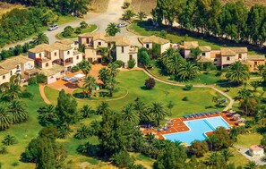 Ett flygbildsfoto av ett semesterområde med flera villor, ett centralt poolområde med solstolar och omgivande grönska.
