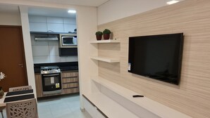 Cocina moderna con microondas, estufa y horno integrados. Hay un televisor montado en la pared, un estante con plantas en macetas y una zona de comedor con una silla y una mesa.