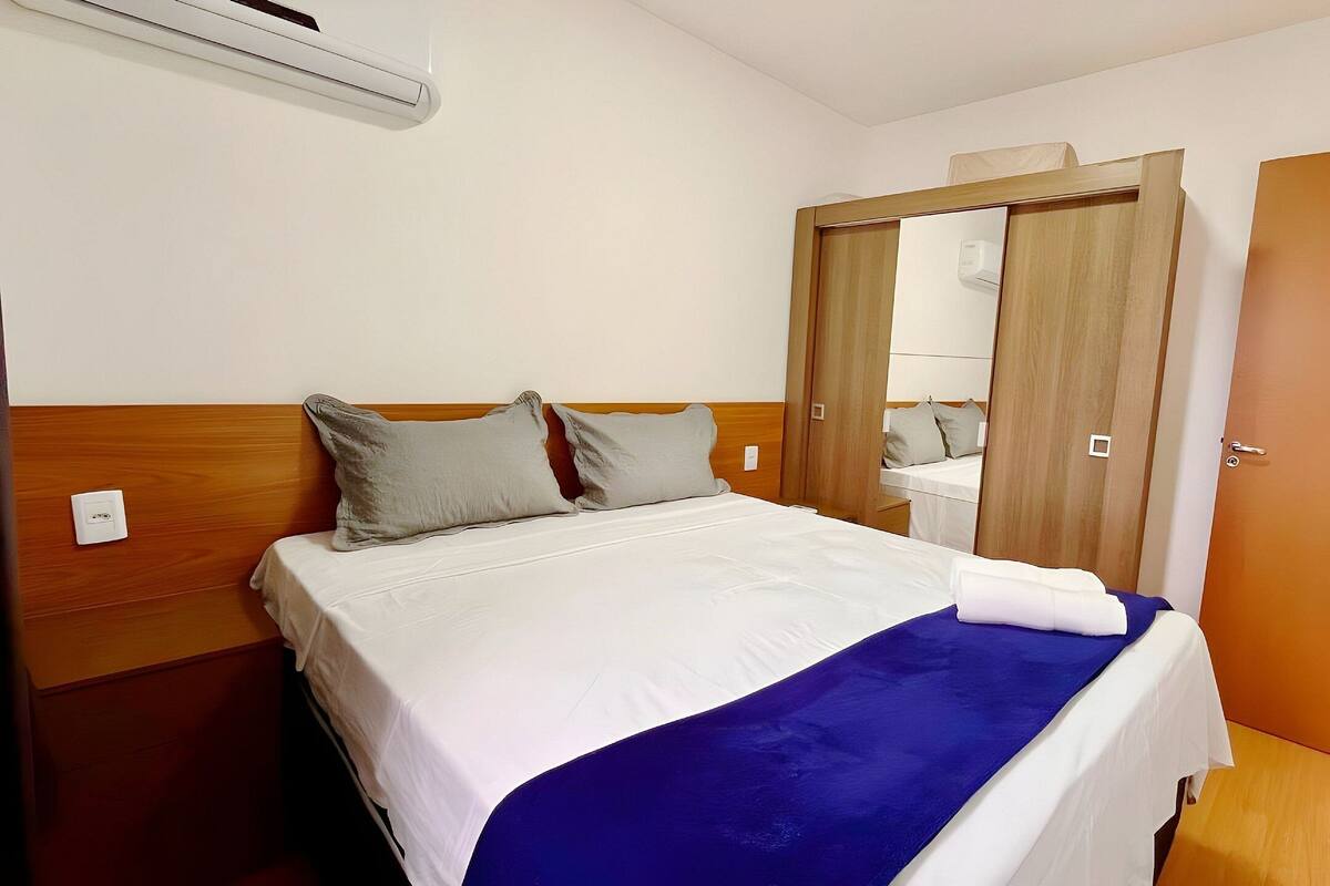 Une chambre d’hôtel avec un lit, une tête de lit en bois, un miroir, une armoire et une salle de bain visible à travers une porte ouverte.