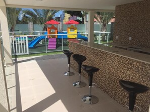 Un patio con un parque infantil, una barra con taburetes y piso de baldosas.
