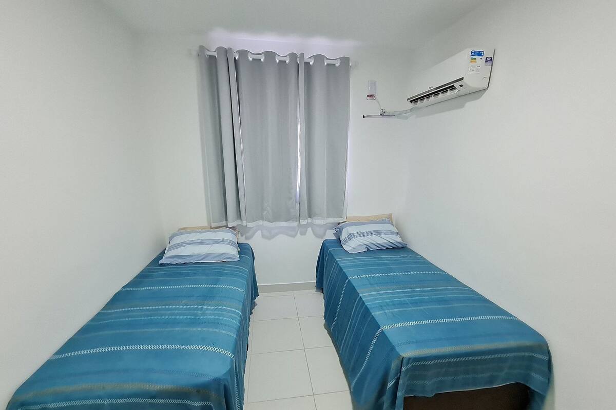 Une petite chambre propre, avec deux lits simples, une fenêtre avec des rideaux, un appareil de climatisation et une étagère fixée au mur.