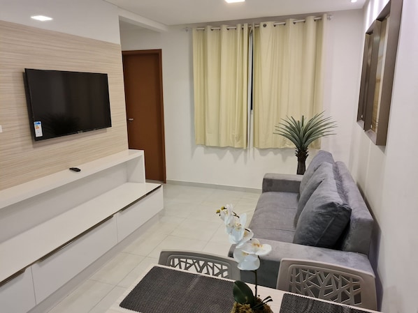 Una sala de estar moderna con un televisor de pantalla plana, un sofá gris, una mesa de comedor con una orquídea decorativa y una planta en maceta.