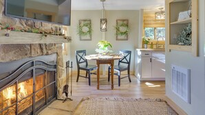 A cozy living room with a fireplace, a dining table with chairs, a TV mounted on a stone wall, and a kitchen area with cabinets and a window. - 1 Mi to Sugar Mountain! Cozy Lodge-Style Condo (Banner Elk)