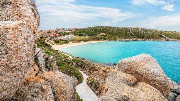 Ein Küstenblick mit klarem türkisblauem Meer, felsigem Gelände und einer kleinen Siedlung mit Gebäuden im Hintergrund.