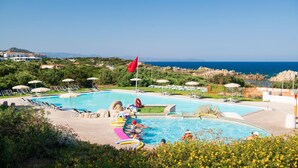 Ein Schwimmbecken mit Liegestühlen und Sonnenschirmen, umgeben von Grünflächen und Blick aufs Meer.