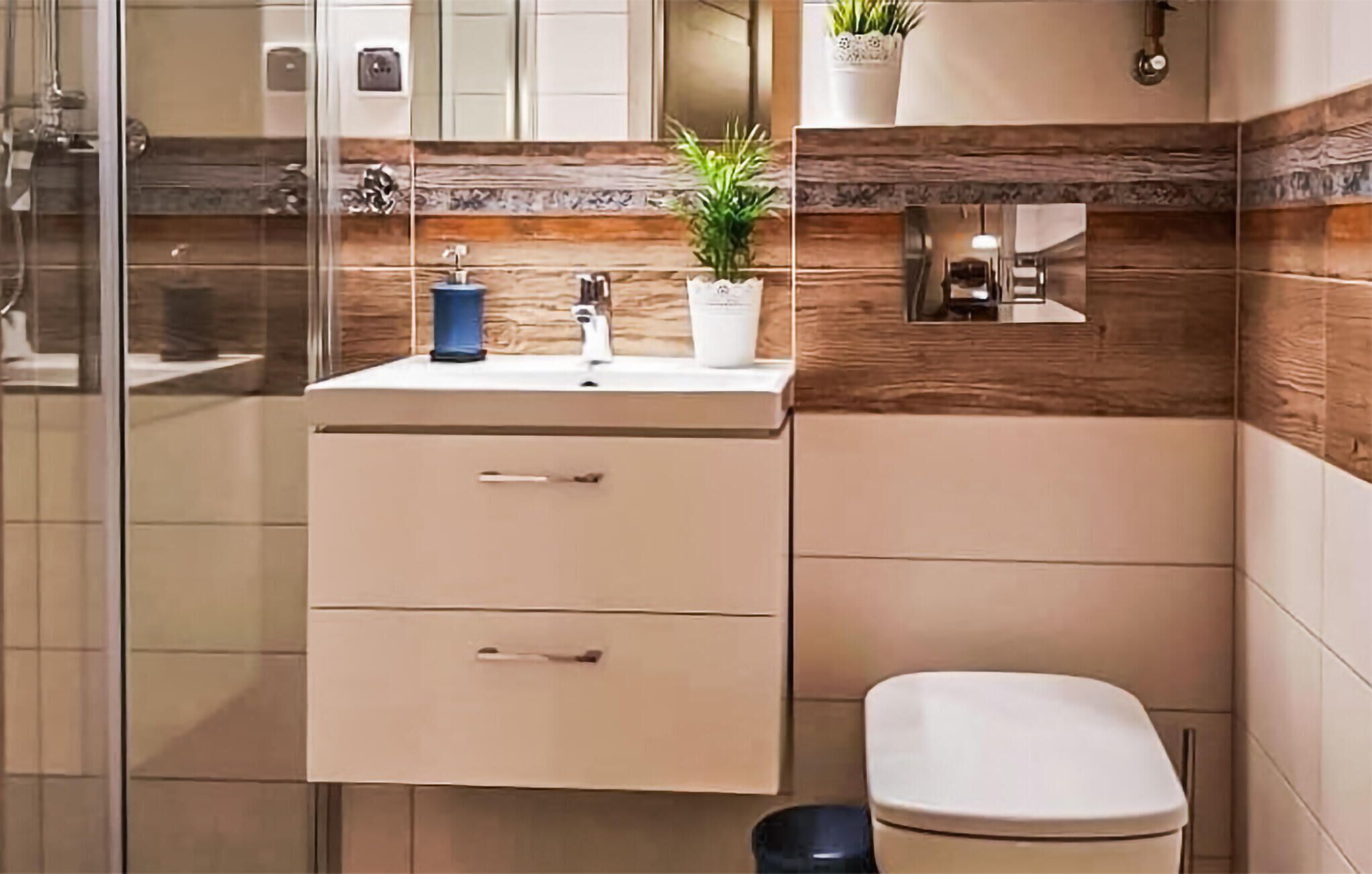 A modern bathroom with a glass shower enclosure, a beige vanity with two drawers, a white toilet, and a small shelf with a potted plant.