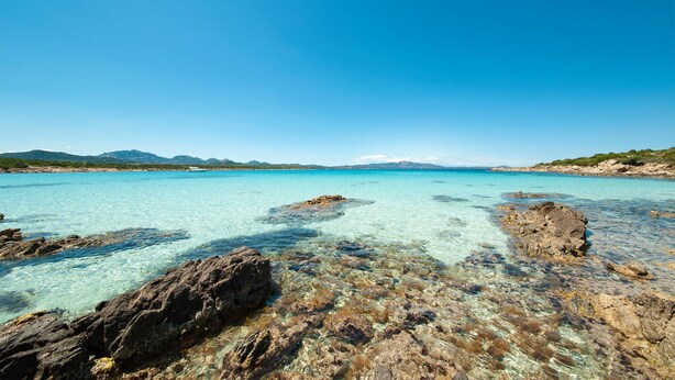 Une vue dégagée sur la côte, avec des rochers au premier plan et des montagnes à l’horizon.