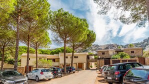 Un espace de stationnement avec plusieurs voitures, une rangée de bâtiments aux murs de pierre et des arbres au premier plan.