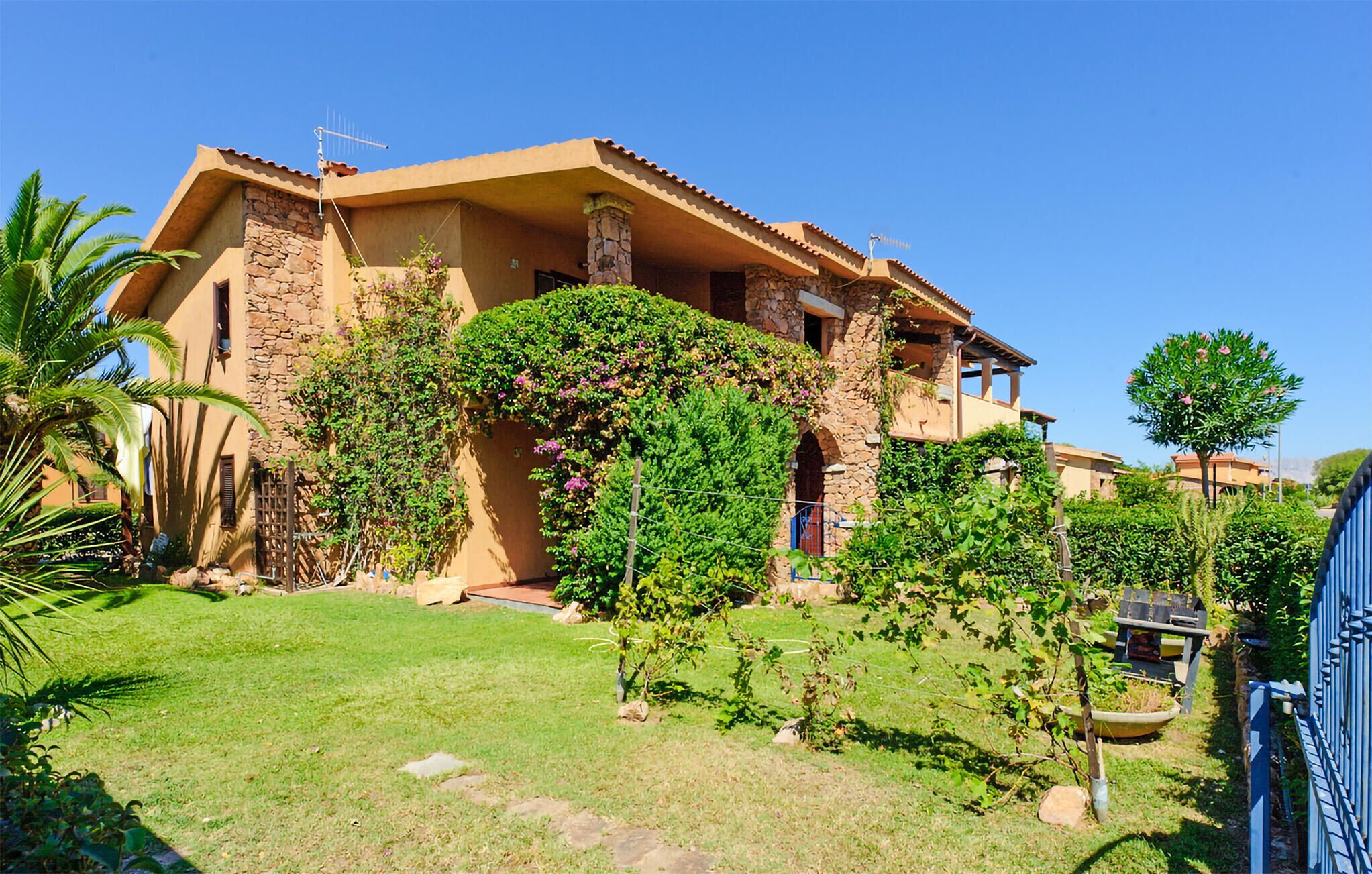 A two-story house with stone walls and a tiled roof, surrounded by a well-maintained garden with various plants and a palm tree.