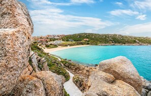 Vista costeira com mar azul-turquesa límpido, terreno rochoso e um pequeno povoado com edifícios ao fundo.
