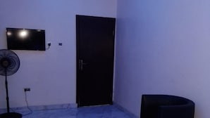 Una habitación con una cama, una silla negra, una mesita, un ventilador de pie y un televisor montado en la pared.