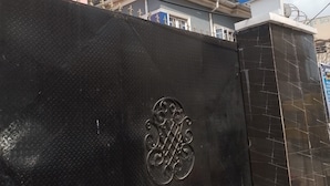 Entrada de hotel con una reja negra que presenta un emblema decorativo, un cartel que indica servicios como alojamiento, restaurante, bar y lavandería, y un edificio rosa vecino con la leyenda "HOTEL".