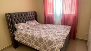 Un dormitorio con cabecera capitonada, colcha estampada y cortinas rojas traslúcidas.