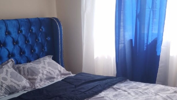 Una cama con cabecera azul, ropa de cama con estampado blanco y gris, y cortinas azules translúcidas.