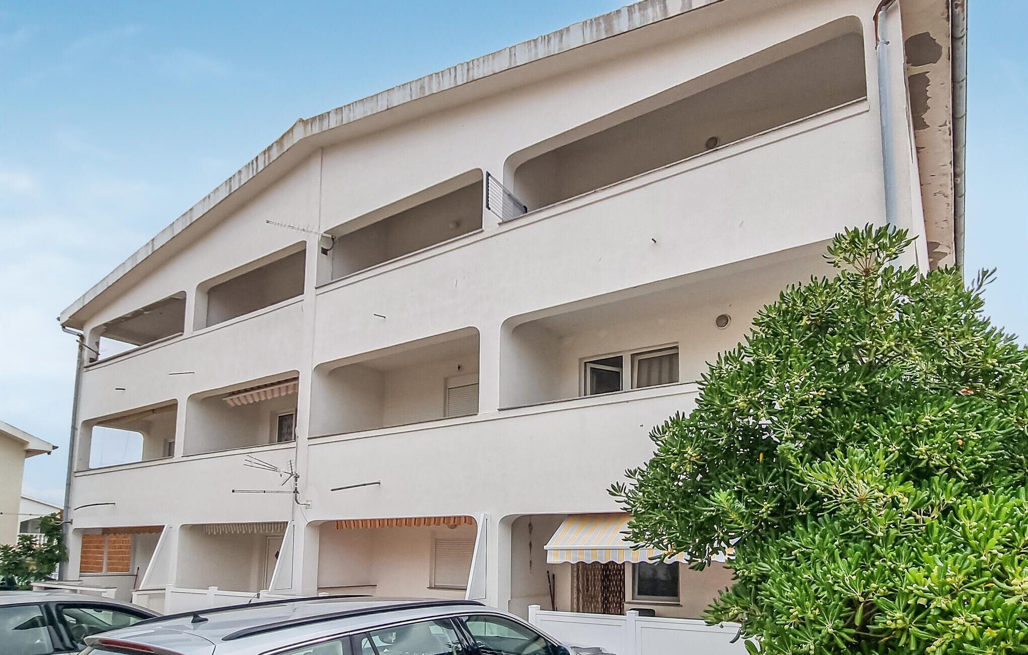 A multi-story building with balconies, a tree in the foreground, and parked cars.