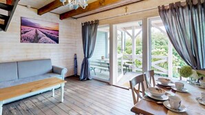 Ein gemütliches Zimmer mit Holzboden, einem grauen Sofa, einem Esstisch mit Tellern und Tassen sowie Blick ins Freie durch große Fenster.