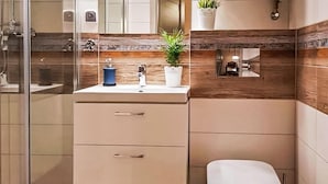 Ett modernt badrum med en duschavdelning med glasväggar, ett beige handfat med två lådor, en vit toalett och en liten hylla med en krukväxt.