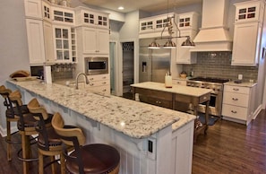 A kitchen with white cabinets, a granite countertop, stainless steel appliances, and a bar area with stools. - NEW Coastal Club Luxury *16117 Rock *4BR Ranch House - Sleeps 10 (Lewes)