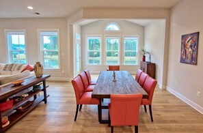 A dining area with a wooden table and red upholstered chairs, large windows, and a painting on the wall.