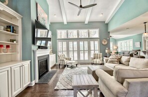 A living room with a fireplace, a ceiling fan, and a large window with shutters.