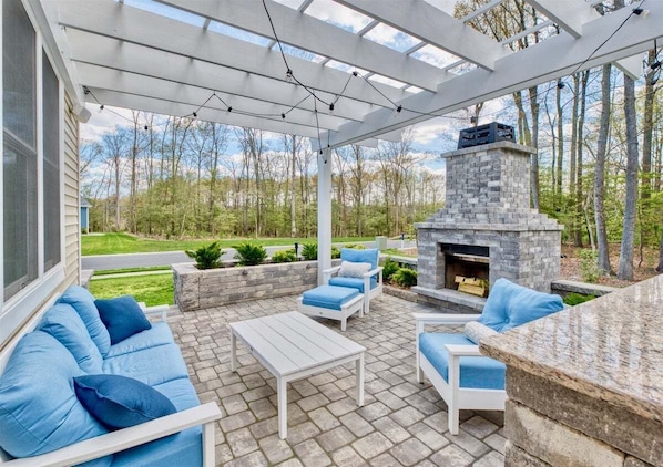 An outdoor patio with a stone fireplace, white furniture, and a pergola.