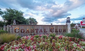 Ein hölzernes Schild mit der Aufschrift 'COASTAL CLUB', umgeben von einem Garten mit verschiedenen Blumen.