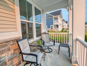 A covered porch with two chairs and a small table, a brick wall, and a view of a two-story house.