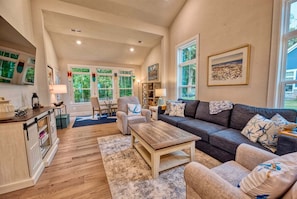 A spacious living room with a sofa, armchairs, a coffee table, and a fireplace. There are large windows with a view of greenery outside.