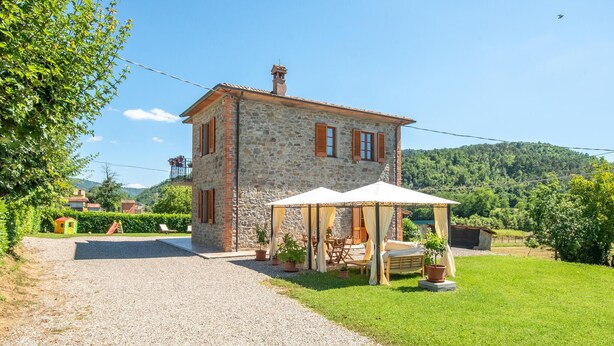 A stone house with a covered patio, a gravel driveway, and a lush green lawn.