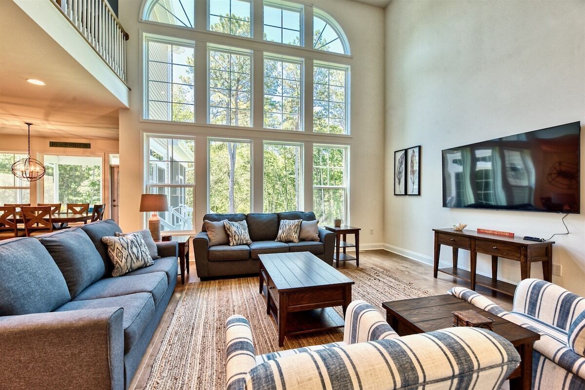 A spacious living room with a high ceiling, large windows, a sofa set, a coffee table, and a flat-screen TV.