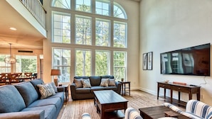 A spacious living room with a high ceiling, large windows, a sofa set, a coffee table, and a flat-screen TV.