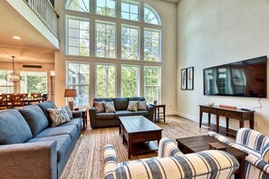 A spacious living room with a high ceiling, large windows, a sofa set, a coffee table, and a flat-screen TV.