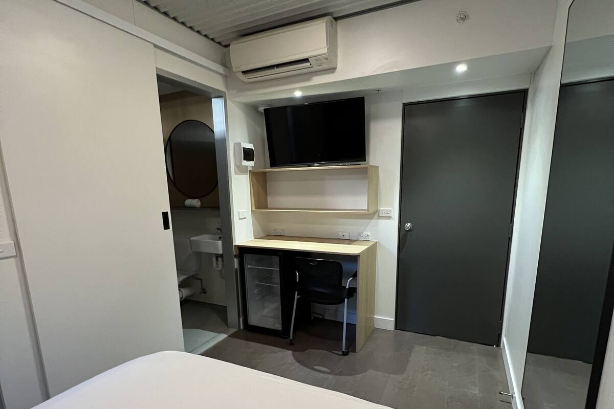 A compact room with a bed, a desk, a chair, a TV mounted on the wall, and a door.
