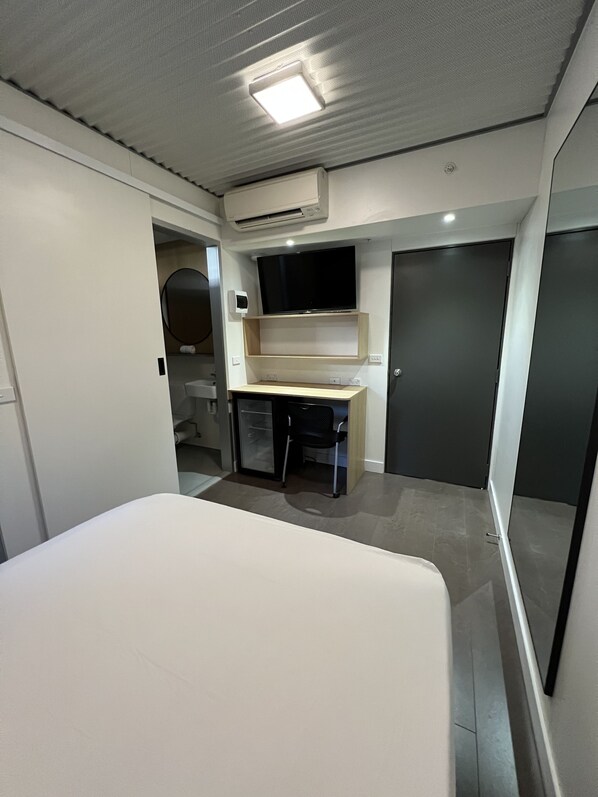 Habitación pequeña con cama, escritorio, silla, televisor montado en la pared y una puerta.