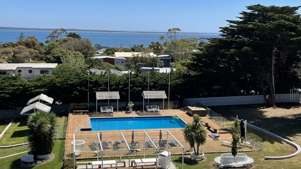 Une piscine entourée d’arbres et de bâtiments, avec des chaises longues et des parasols à proximité.