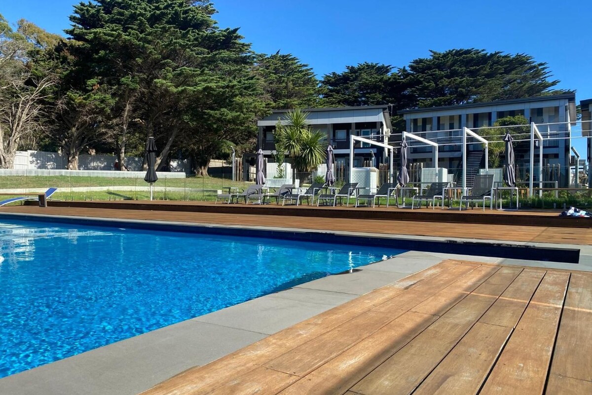 Una piscina con cubierta de madera, sillones de descanso y un edificio al fondo.