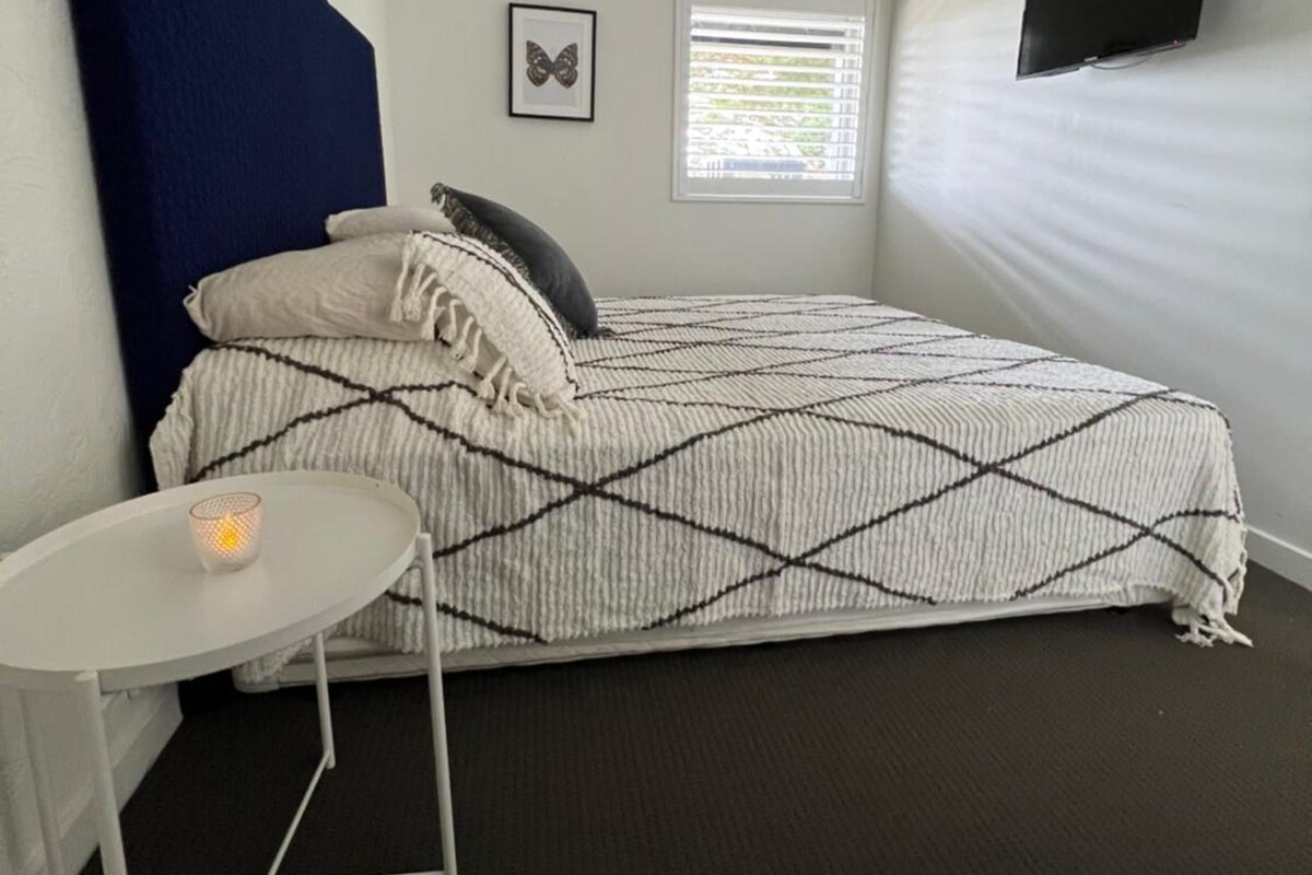 Une chambre à coucher avec un lit, une petite table sur laquelle est posée une bougie, une fenêtre avec des stores et un téléviseur fixé au mur.