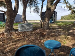 A rustic outdoor seating area with metal barrels and a blue table, surrounded by trees and a building in the background. - Dog friendly! - #13 is 1 of 3 containers now dog friendly! (Blanco County)